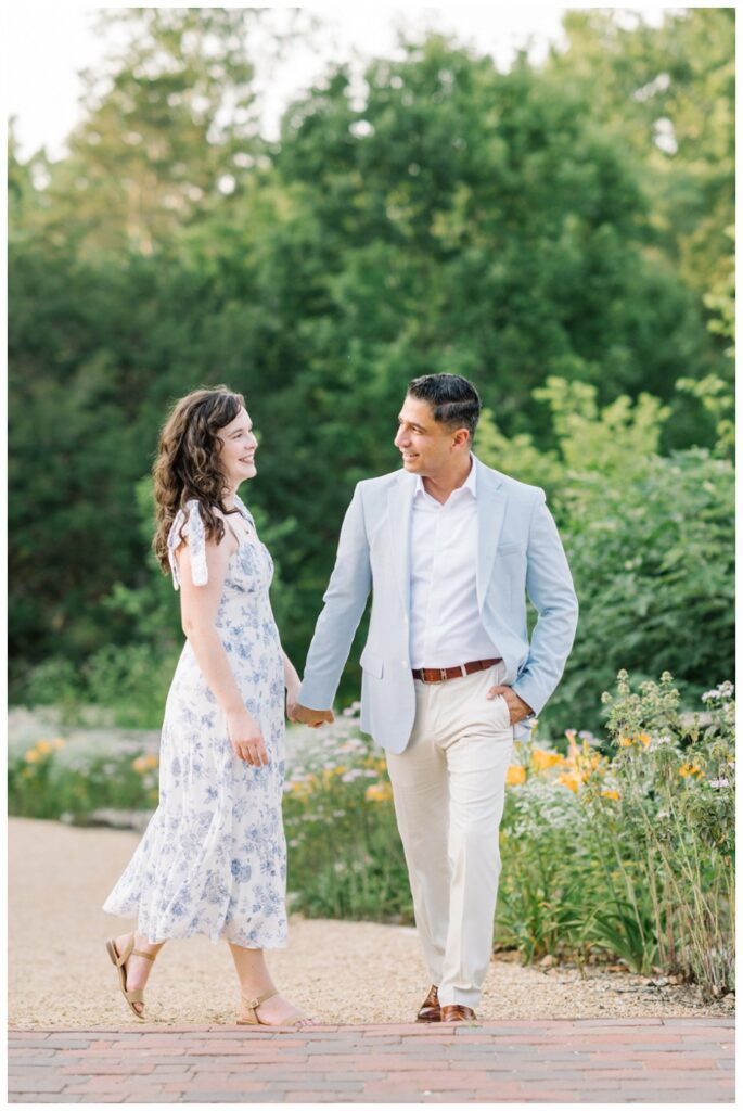 Stylish garden engagement session in Colonial Williamsburg. Man in a powder blue sport coat and woman in a white dress with a blue floral pattern and bows on the straps.