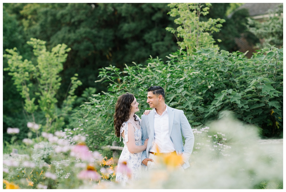 Stylish garden engagement session in Colonial Williamsburg. Man in a powder blue sport coat and woman in a white dress with a blue floral pattern and bows on the straps.